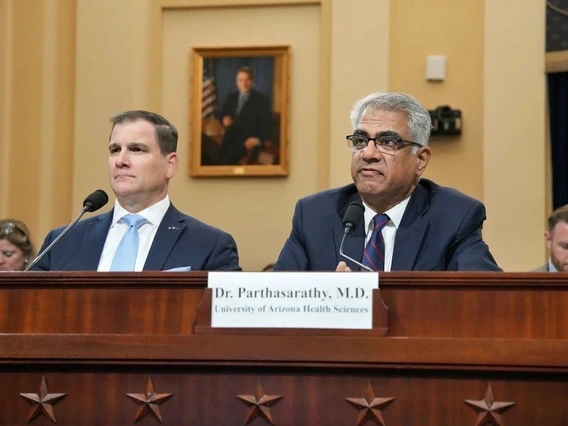 Dr. Parthasarathy, M.D., from the University of Arizona Health Sciences, testifies at a congressional hearing, seated at a wooden desk with a nameplate in front of him. He wears a dark suit, glasses, and a striped tie, speaking into a microphone beside another suited man. The background shows a formal hearing room with a portrait, panelists, and U.S. government decor.