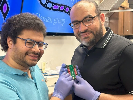 Two smiling researchers in a lab, both wearing glasses and gloves, hold up a small experimental circuit board labeled 'Graphene.' Behind them, a screen displays the words 'Hassan group' along with colorful molecular imagery. The setting suggests a breakthrough in advanced electronics or transistor technology.