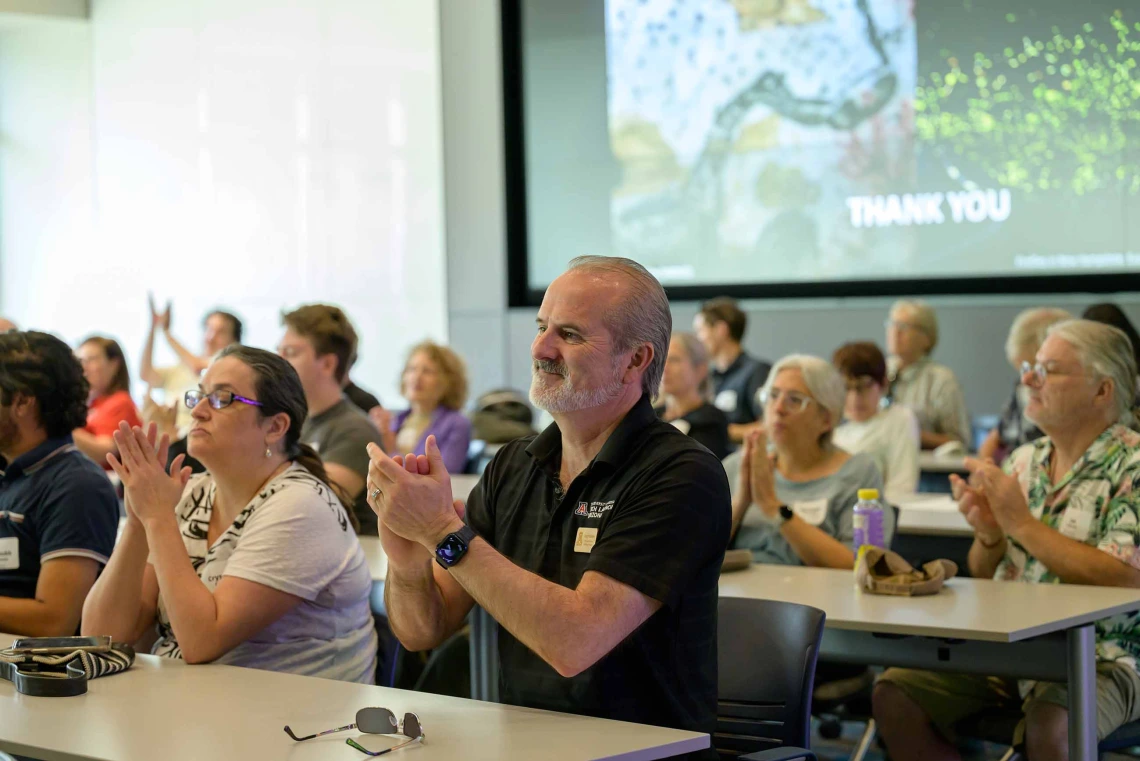 An audience sitting at rows of tables applauds. 
