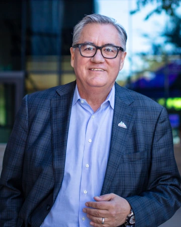 Photograph of Dr. Diaz de la Rubia in a gray plaid suit jacket, standing outside, smiling in a casual pose